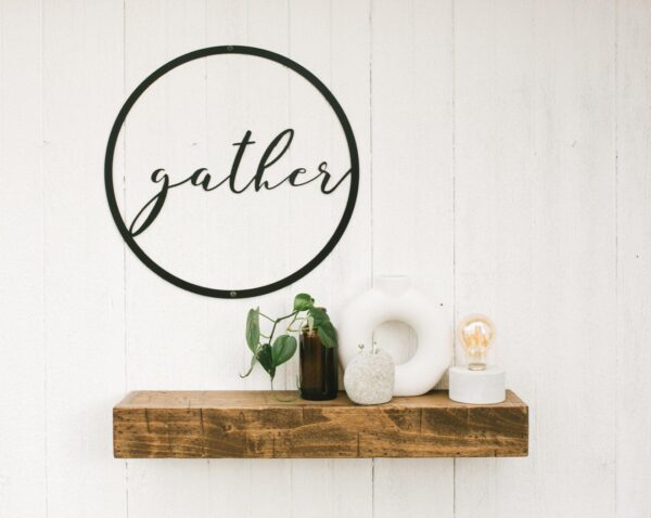 round metal sign with the word gather in the middle - hanging over a small wooden shelf
